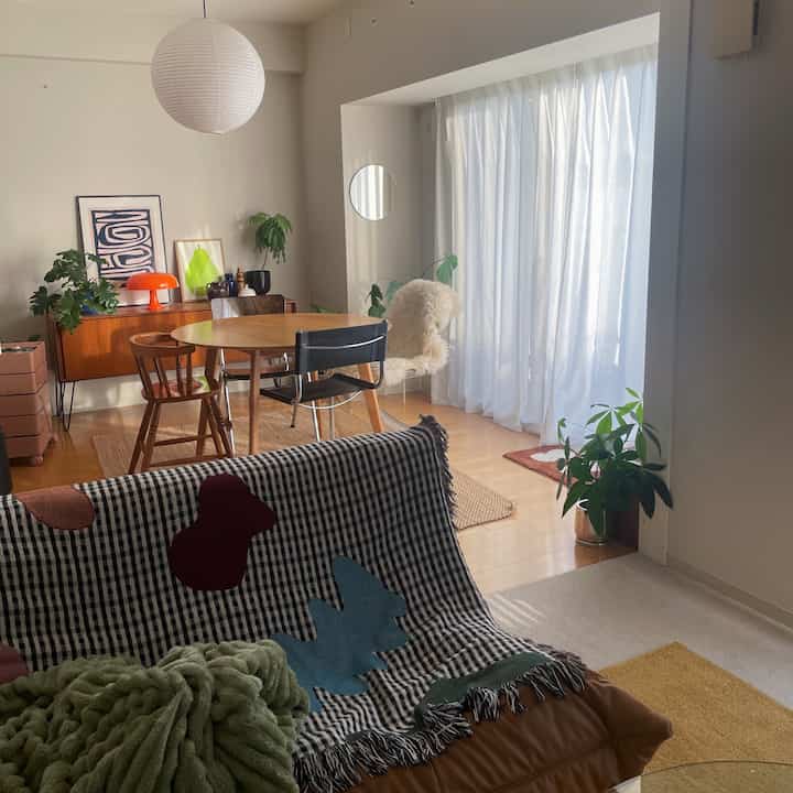 白いカーテンとブラウン家具が配されたナチュラルな色調のリビング、ダイニングテーブルとソファの上のスローが温かみのある空間を演出