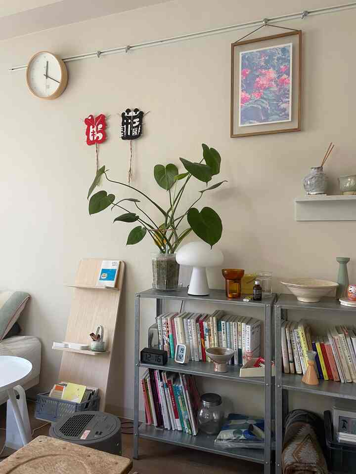 ナチュラルカラーのワンルームリビング、金属製の本棚に本が並び、白いテーブルランプと植物が置かれた落ち着いたインテリア