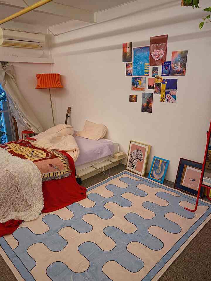 ピンクと赤のアクセントがあるワンルームの寝室、パズル柄のカーペットと壁に配置されたポスターが印象的な雰囲気のあるインテリア