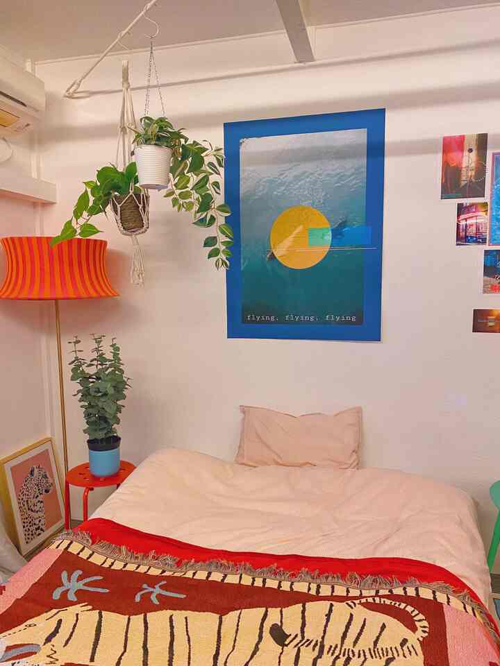 ピンクの寝具とアートポスターが飾られた小さなワンルーム寝室、植物とオレンジ色の照明がアクセントの自然な雰囲気のインテリア