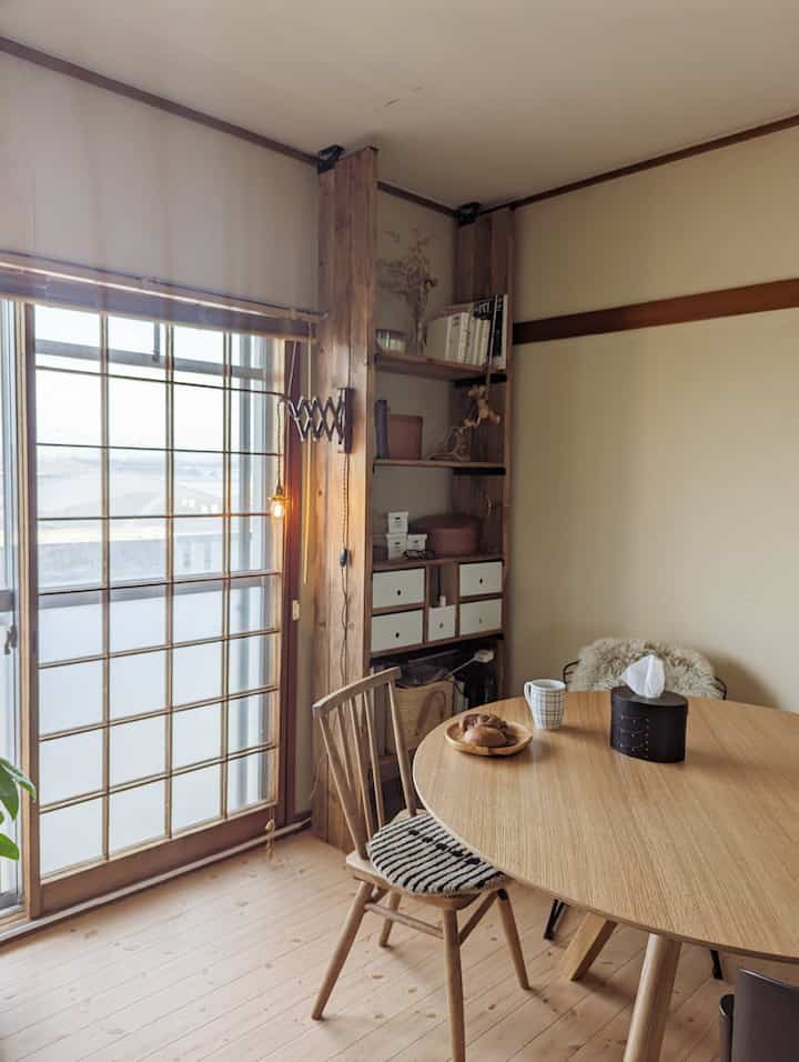 Natural tone dining room featuring a round wooden table, wooden chair, wall-mounted shelves, and wall sconce with cozy atmosphere