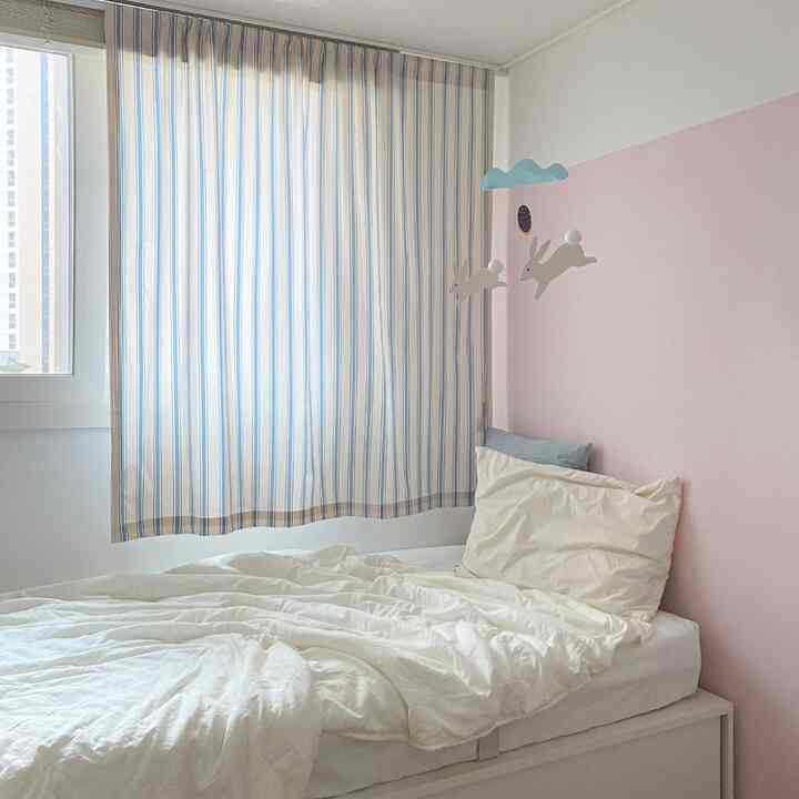 ピンクの壁と青いストライプのカーテンが調和した子供部屋、白い寝具が温かみのある雰囲気を演出。