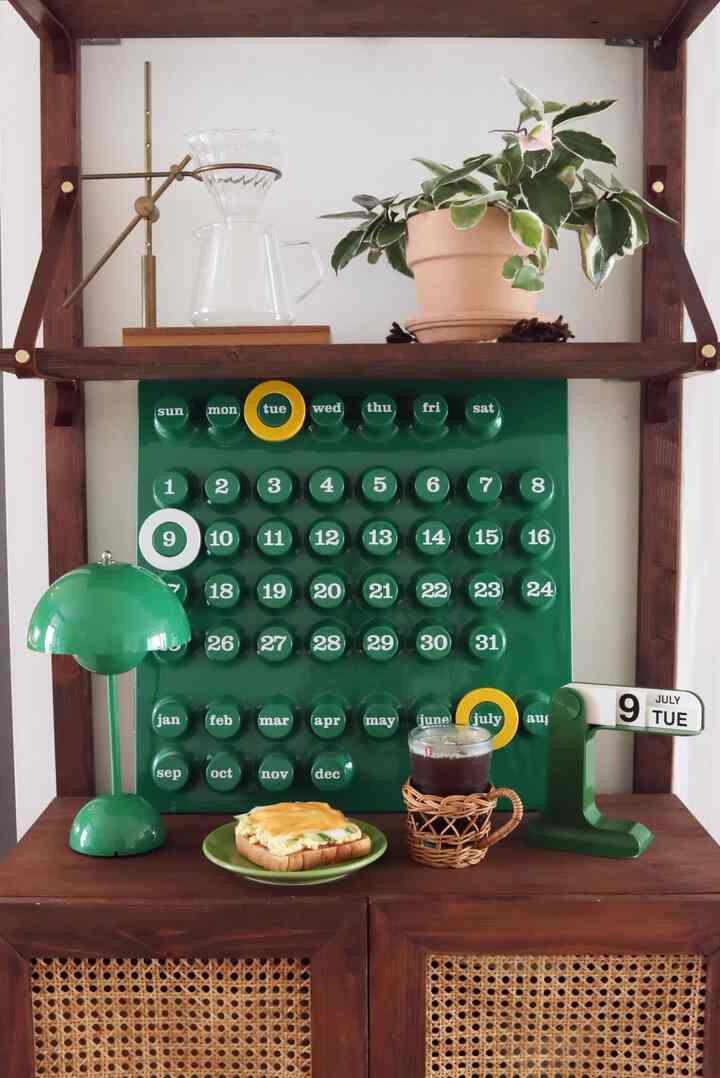 Cozy home cafe space featuring a green calendar, wooden shelf with plant and coffee accessories, and green-toned lighting and tabletop items