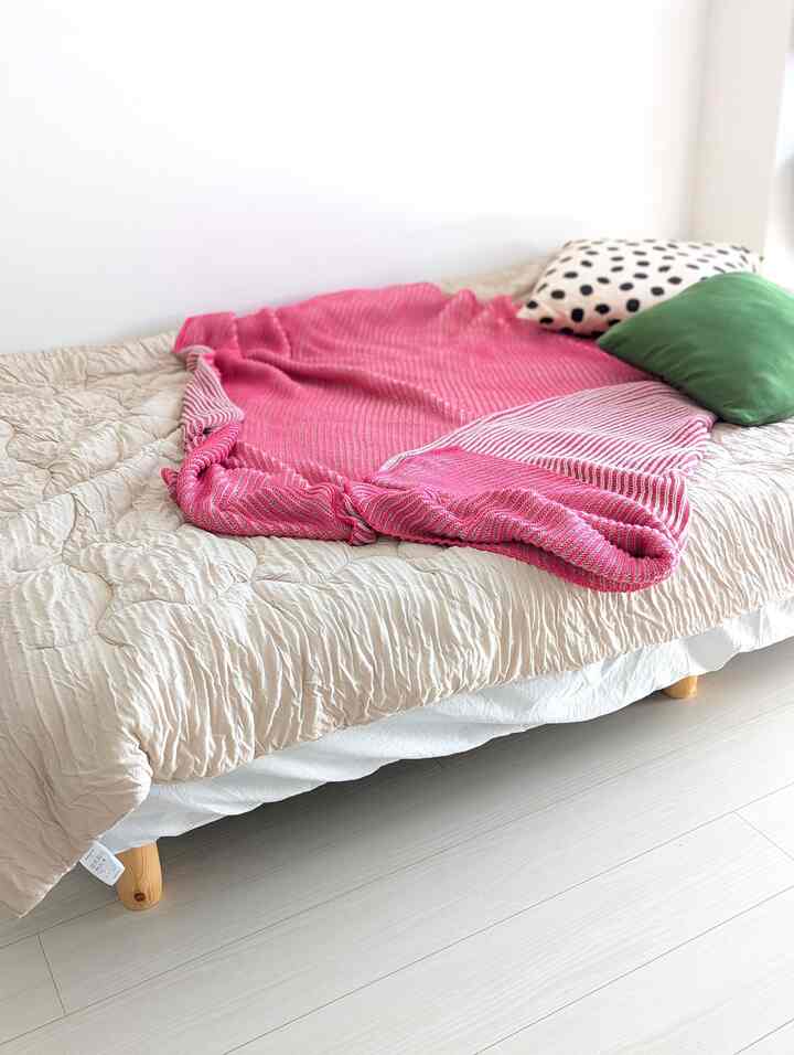 ベージュとピンクがアクセントの寝室、ナチュラルな木製脚のベッドとクッションが配置されたシンプルで落ち着いた印象のインテリア