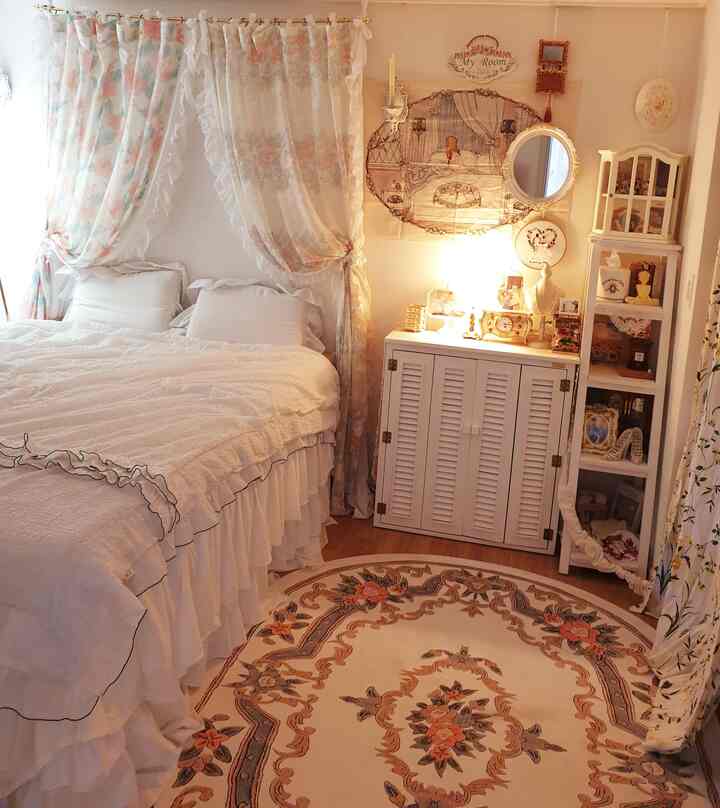 白とヴィンテージ調の寝室、レースのフリルカーテンと花柄の楕円ラグ、アンティーク風家具が配置された雰囲気のあるインテリア