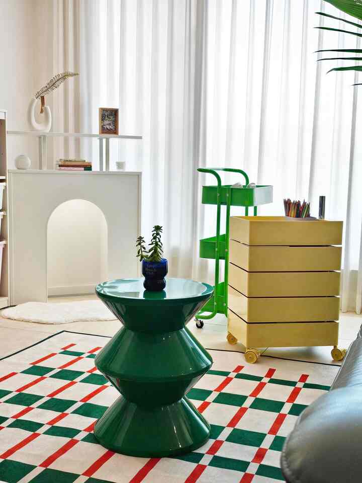 White walls and curtains frame a mid-century modern living room featuring a vivid green stool and colorful trolley carts in a bright setting
