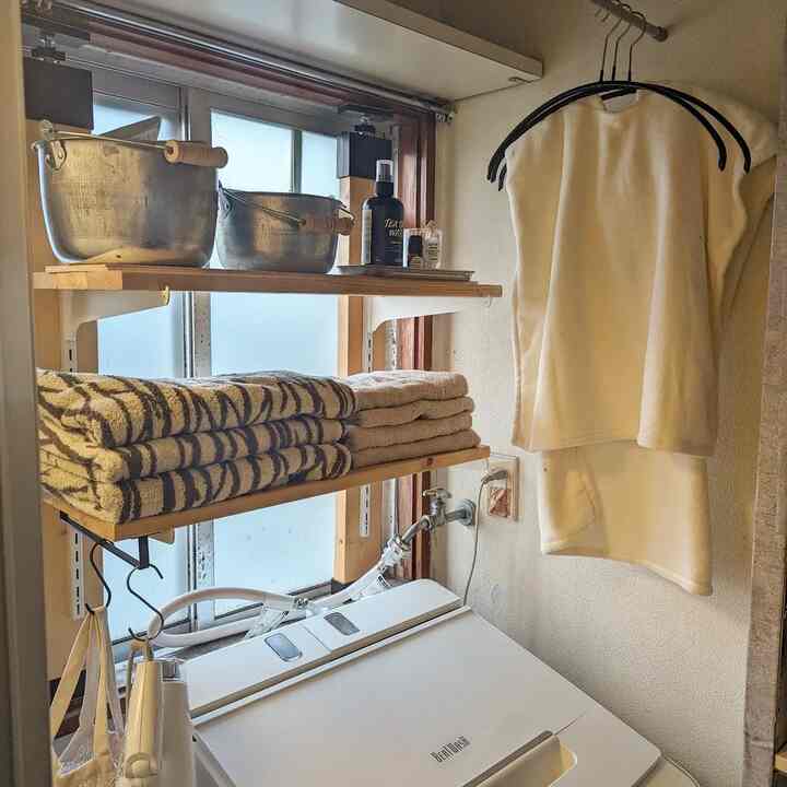 白と木目調が基調の狭い洗濯室。洗濯機の上の棚にはタオルと金属製のバケツが整然と並び、壁には白いバスローブが掛けられた実用的な空間。