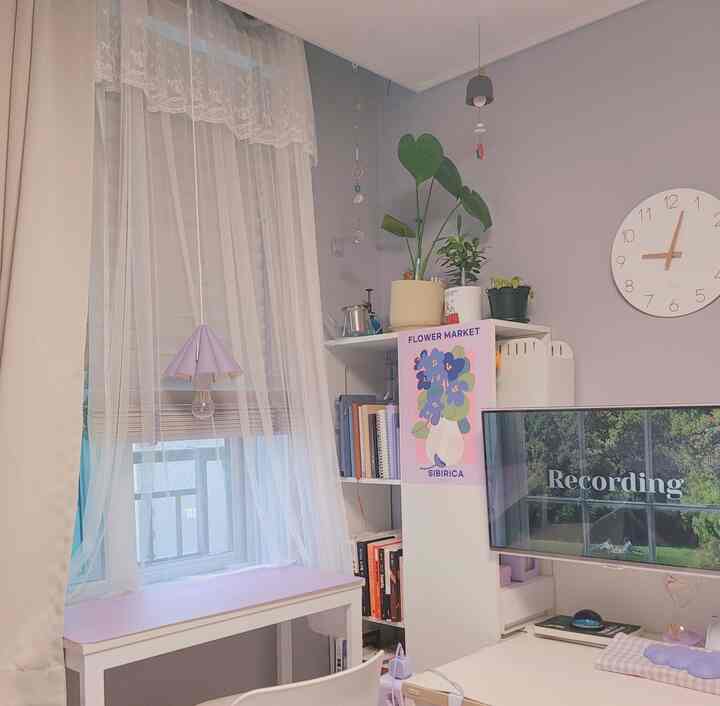 ラベンダー色調の部屋、白いデスクと両面本棚、カーテンと壁掛け時計が配置された落ち着いた雰囲気のインテリア
