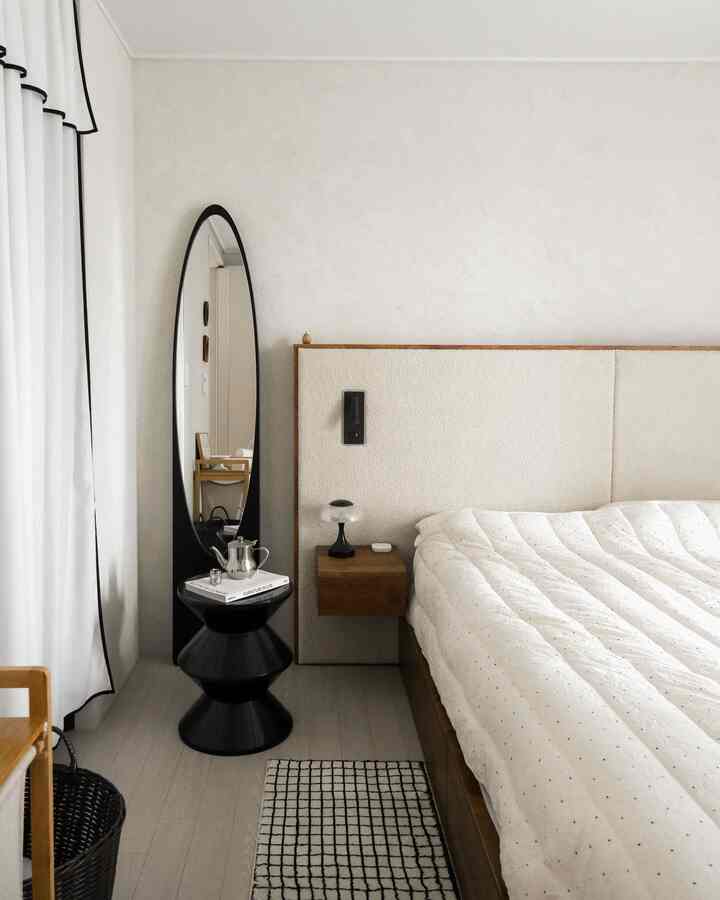 A bedroom with white, black, and wood tones featuring a wooden bed frame, large standing mirror, and black stool in a modern natural interior.