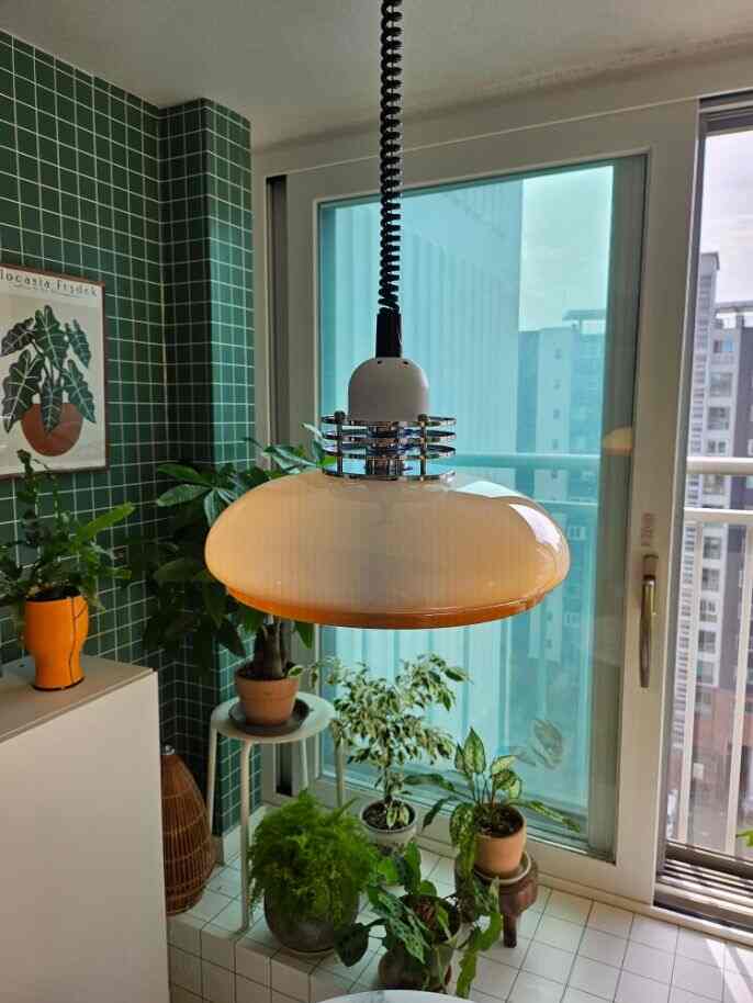 グリーンタイルの壁と多様な植物が配置されたバルコニー窓際のモダンな空間、中央に白いペンダントライトが吊るされている