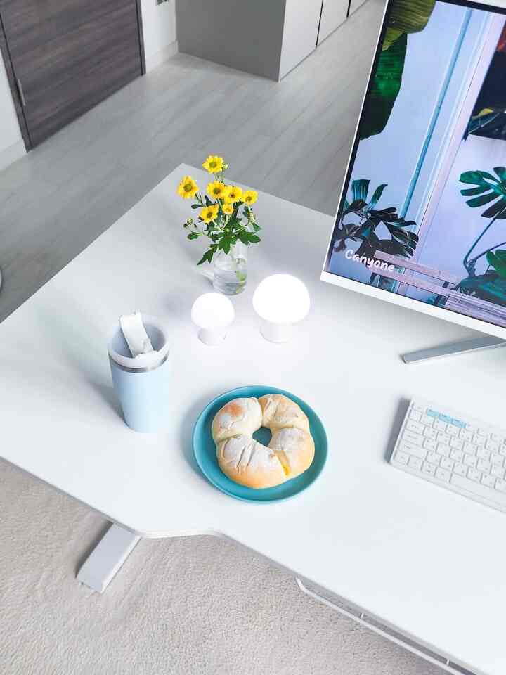 ホワイトとブルーのトーンのホームオフィス、電動スタンディングデスクにキーボードとミニLEDランプ、花とパンが置かれた清潔感のある作業スペース