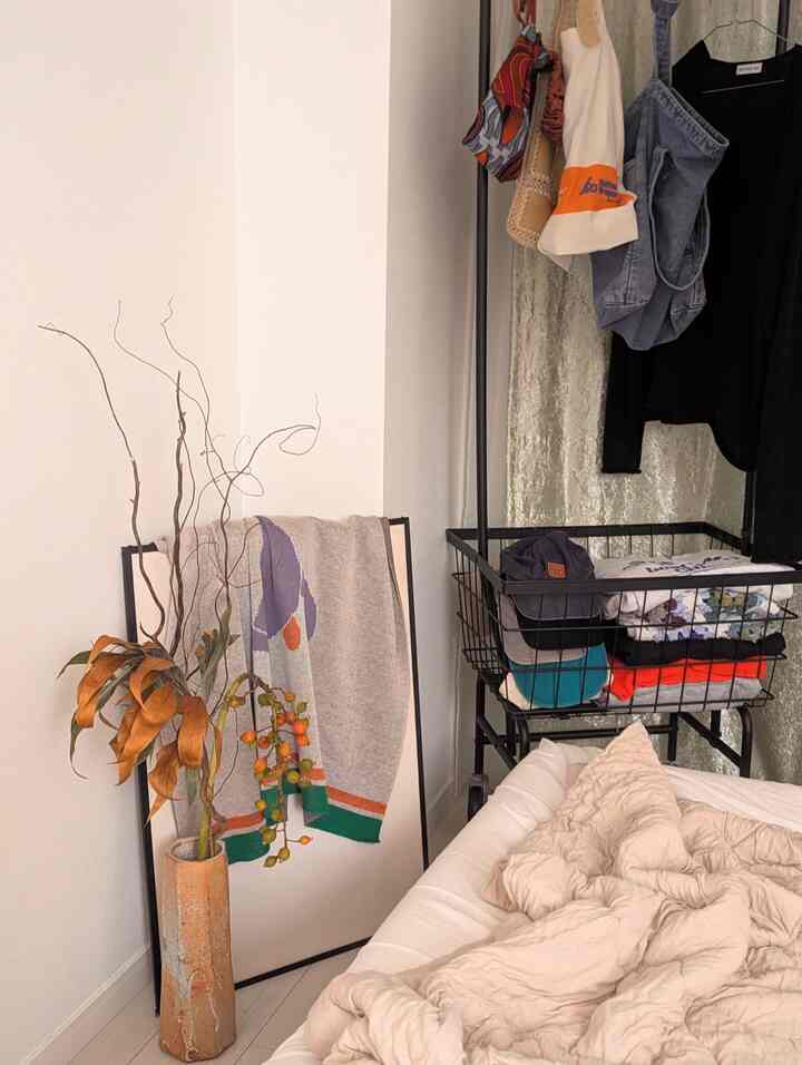 白い壁とベッドが背景の空間に、ポスターにかけられたアートブランケットと茶色のドライプラントが配置された温かみのある秋のインテリア