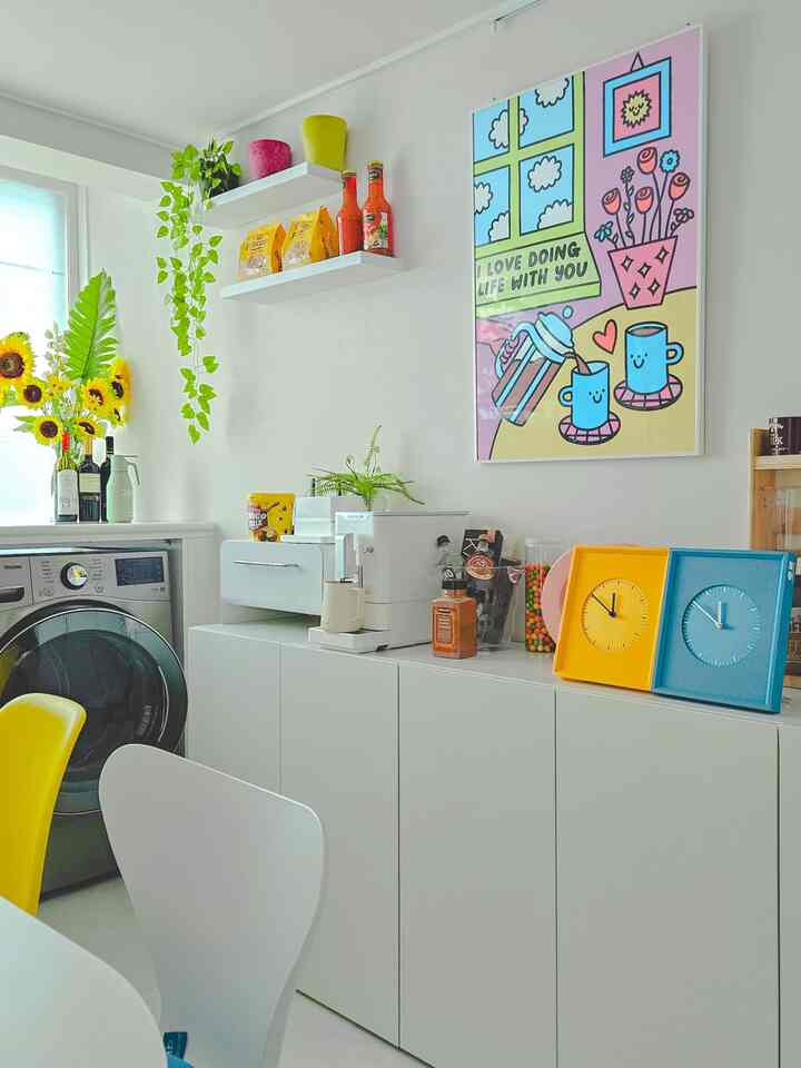 明るい白色調のキッチン、白いキャビネットの上にコーヒーマシンとカラフルな装飾が配置され、活気のある空間