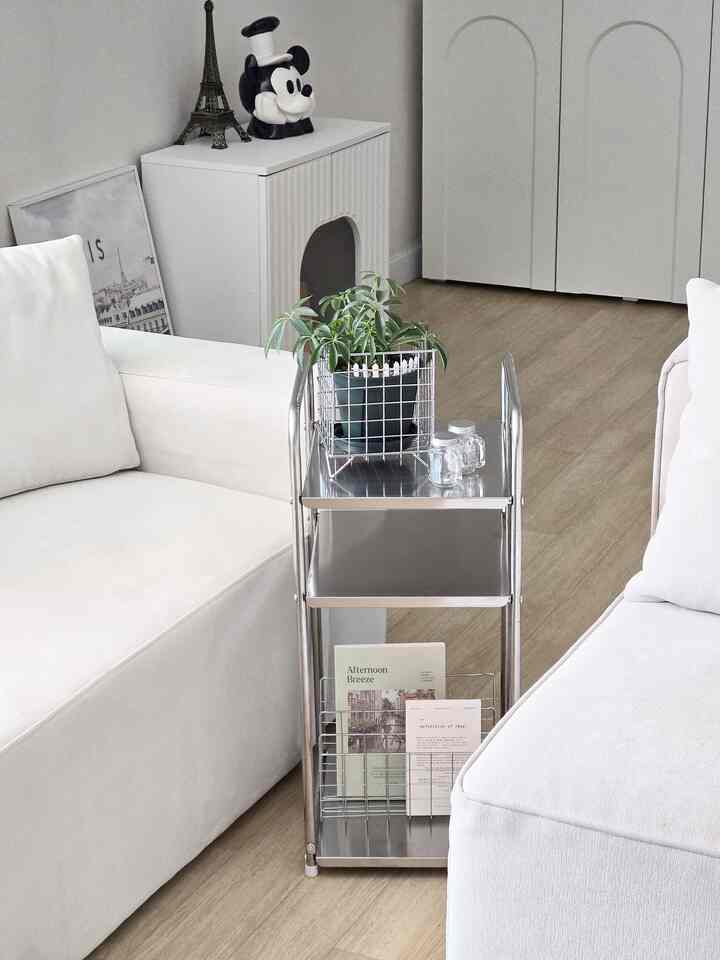 A clean and minimalistic living room dominated by white and silver tones, featuring a sectional sofa, stainless steel shelving, and decorative objets