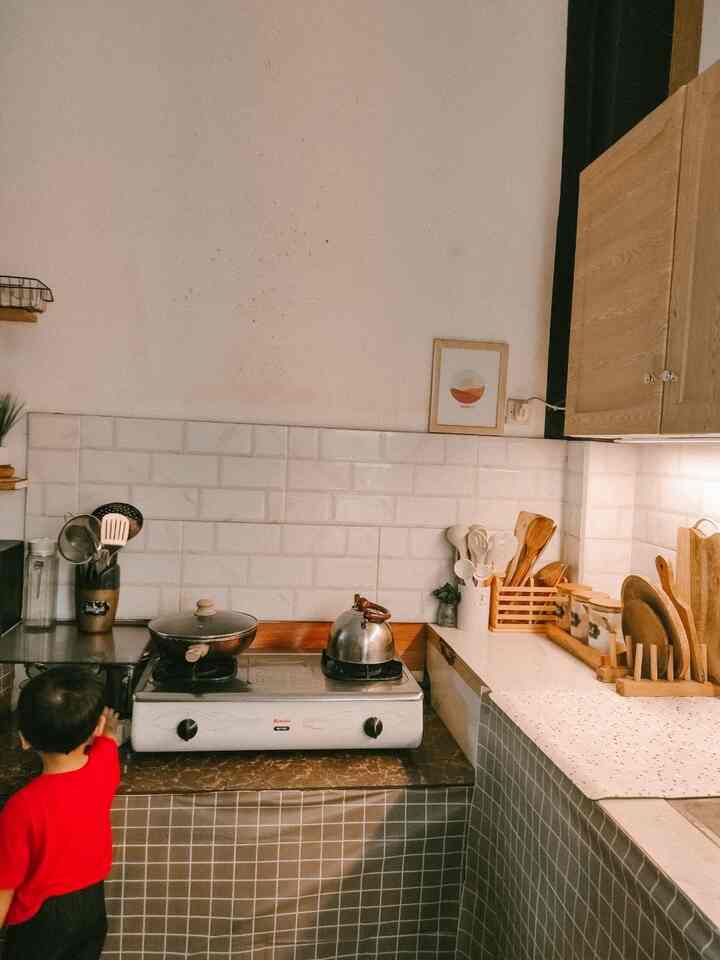 Compact kitchen with white tiled wall, brown kitchen utensils, and a child in red shirt seen near stovetop