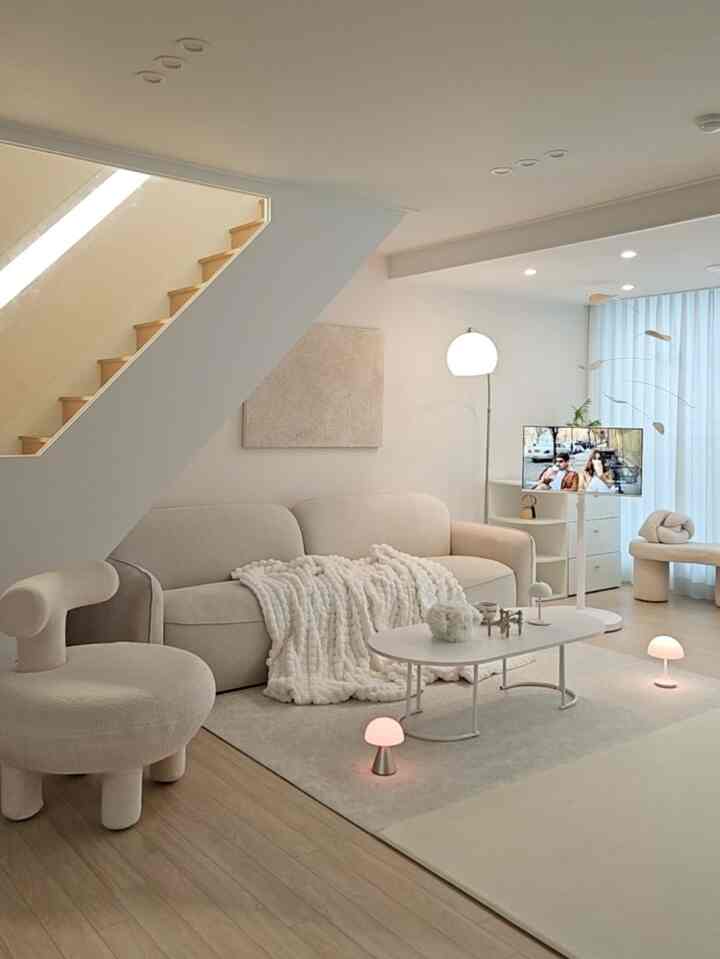 明るいベージュ色調のリビング、柔らかなソファと暖かいブランケット、無線照明と階段が配置された自然で落ち着いた雰囲気のインテリア