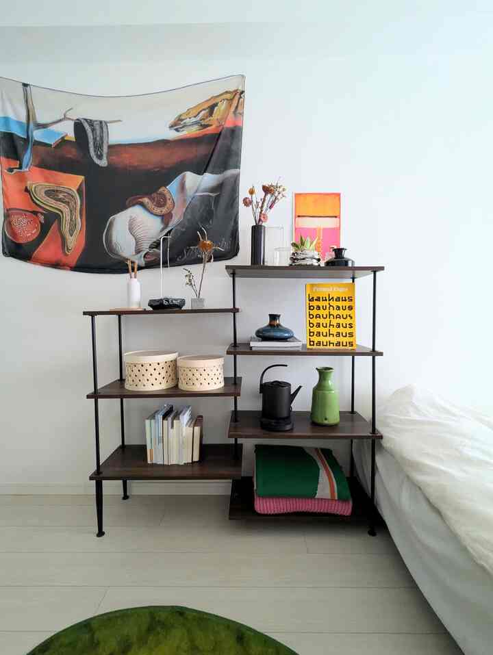 白い壁とベッドのあるナチュラルな寝室、木目調の棚とファブリックポスター、プラントスタンドが配置された清潔感のある空間