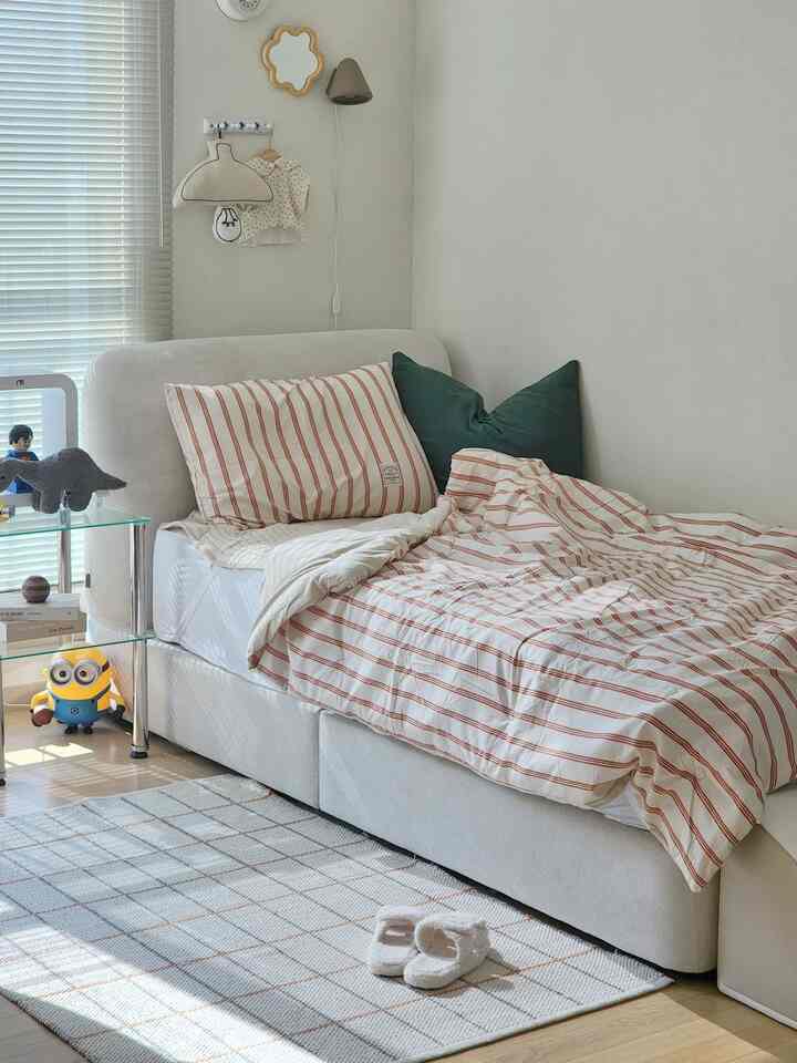 暖かいベージュとホワイトのトーンの子供部屋、ストライプの寝具と緑青色のクッションが配置され、チェック柄のラグが調和した印象的な空間