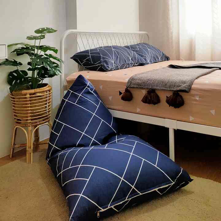 A cozy bedroom with navy and natural colors featuring a bed, a bean bag, and a leafy plant in rattan stand