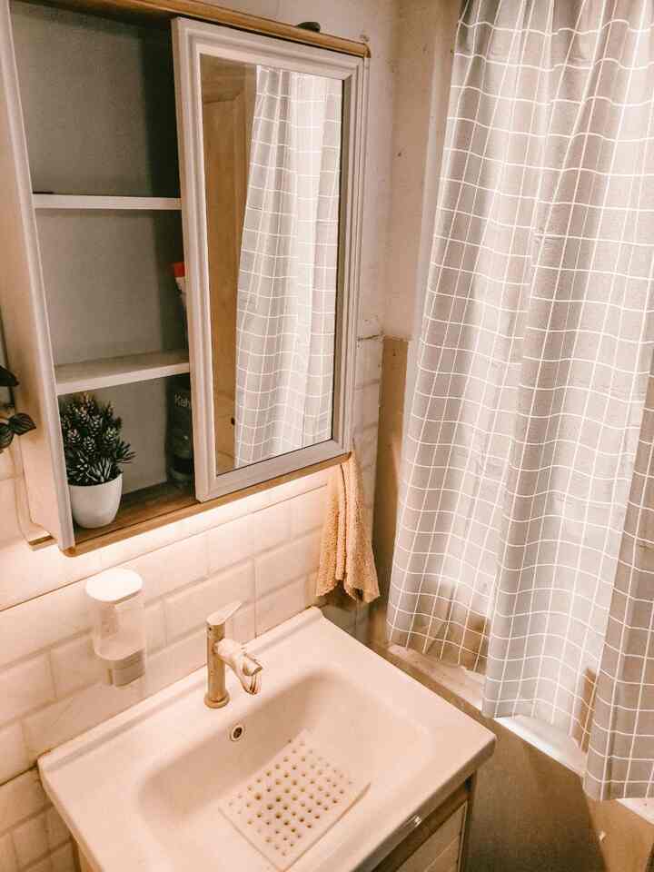 Small bathroom space with white and natural tones, featuring a storage mirror cabinet and curtain over a minimalist washbasin area