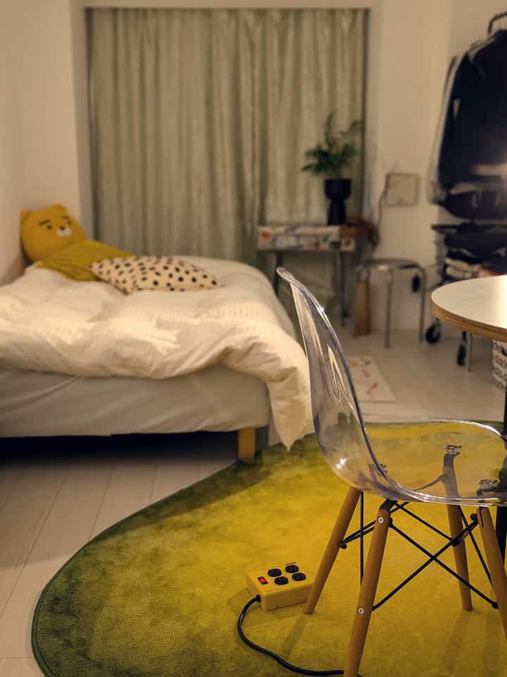 黄色と緑のトーンの小さな寝室、透明な椅子と観葉植物、グラデーションラグが配置された雰囲気のあるインテリア