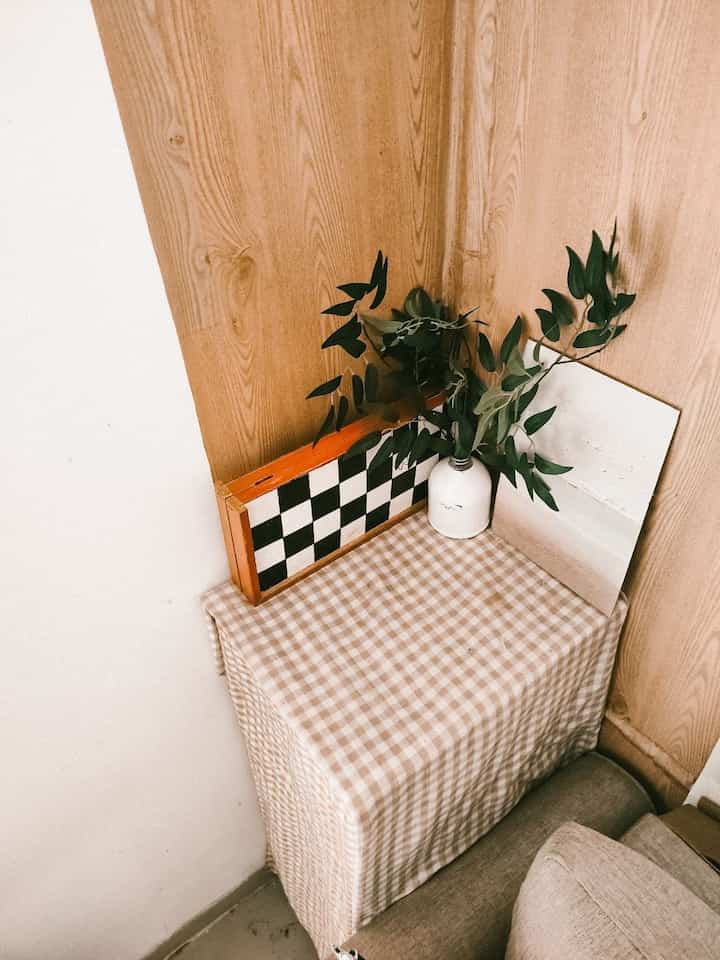 A beige and wood-toned corner of the living room with a checkered cloth-covered storage unit and green artificial leafy decoration creating a simple space
