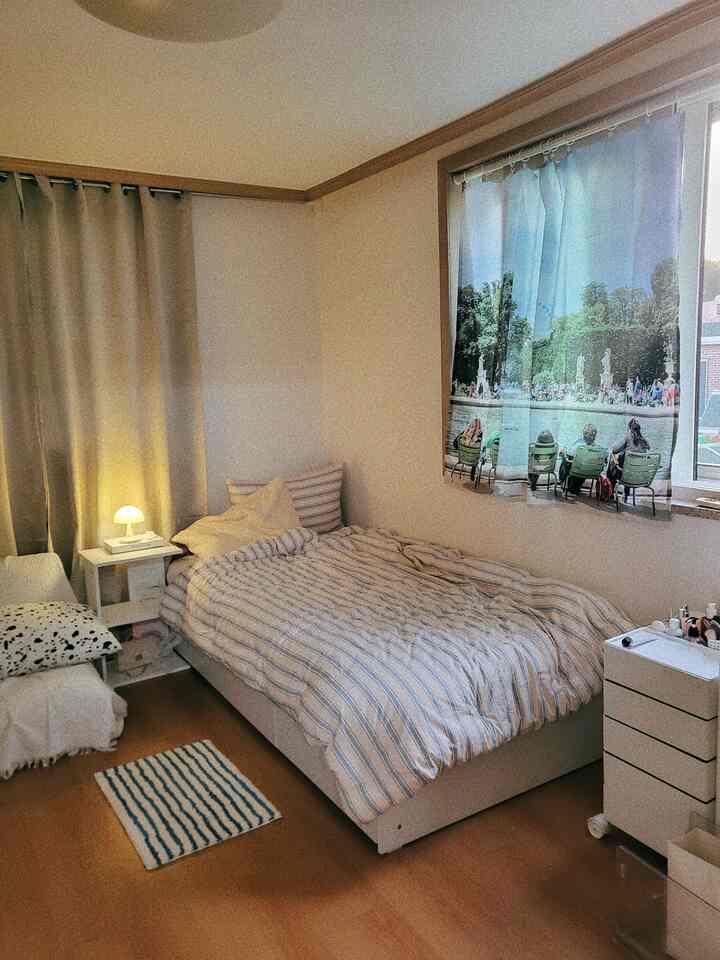 ベージュの壁と白いベッドがある寝室、ストライプの寝具と遮光カーテン、ファブリックポスターが配された落ち着いた空間