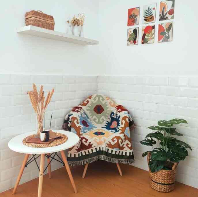 Natural-toned corner space with a white tiled wall, wood floor featuring an armchair draped with colorful patterned throw, a small round coffee table, and a Monstera plant