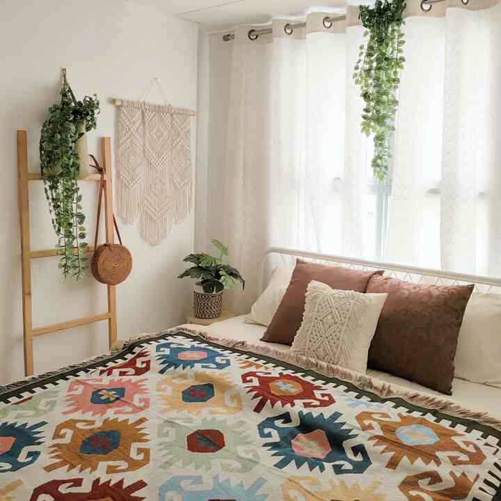 Bright white and beige bedroom featuring plants, wood tone accents, and a colorful Bohemian-style blanket creating a cozy atmosphere