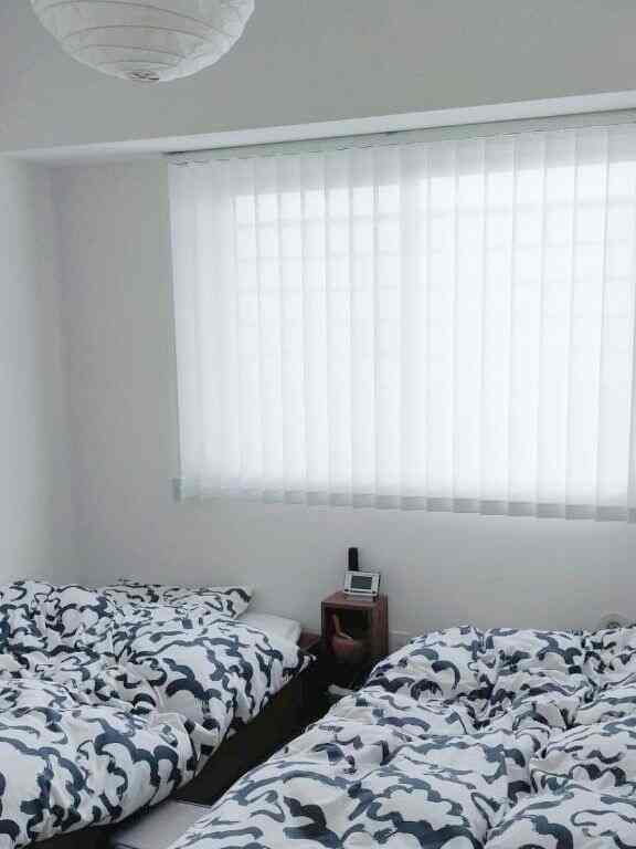 白を基調とした明るい寝室、2台の柄入り掛け布団が配置され、窓には縦型ブラインドが設置されたコンパクトな空間