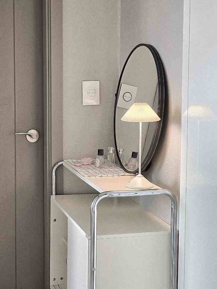 グレーと白のトーンのバスルーム入口、ミッドセンチュリーモダンスタイルのスライディングキャビネットと丸い鏡、白いスタンドランプが配置された印象的な空間