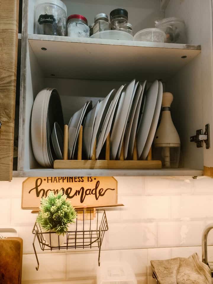 A modern kitchen interior in white and wood tones featuring a cabinet with neatly arranged plates and cooking utensils