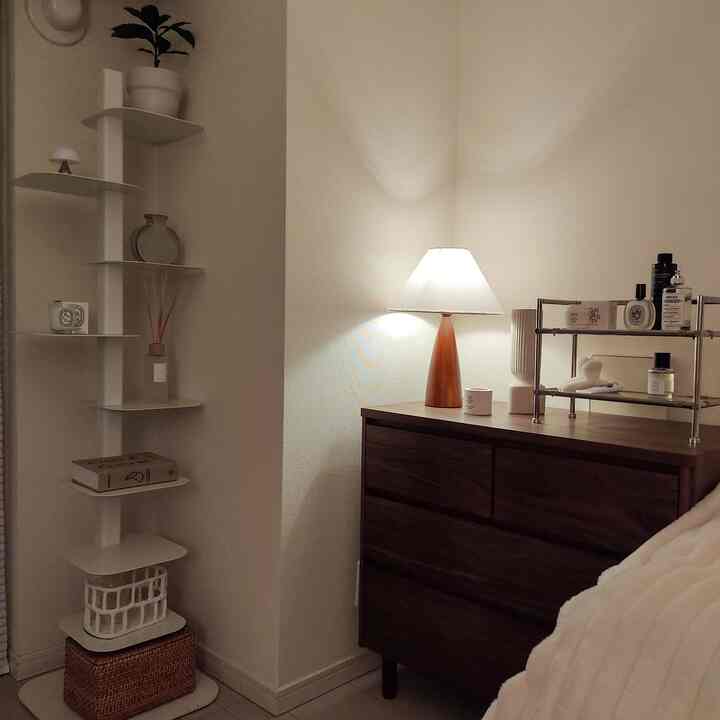 White and natural brown toned 1K bedroom for single household featuring chest of drawers, bookshelf tower, and table lamp in cozy setting