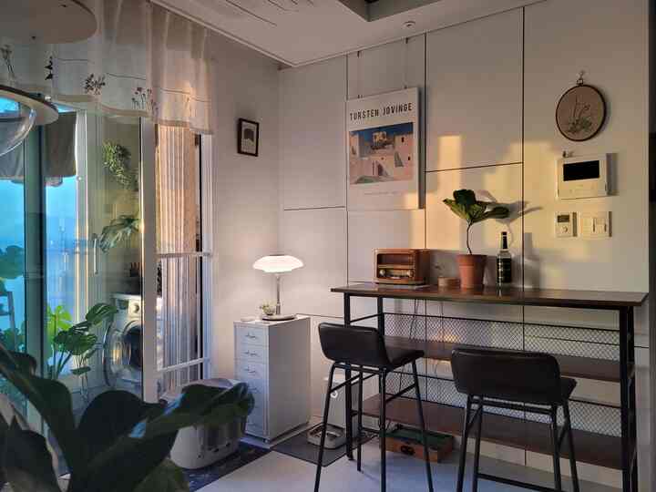 Warm sunlight fills a white-toned home bar space beside a balcony, featuring bar stools and plants for a cozy interior.