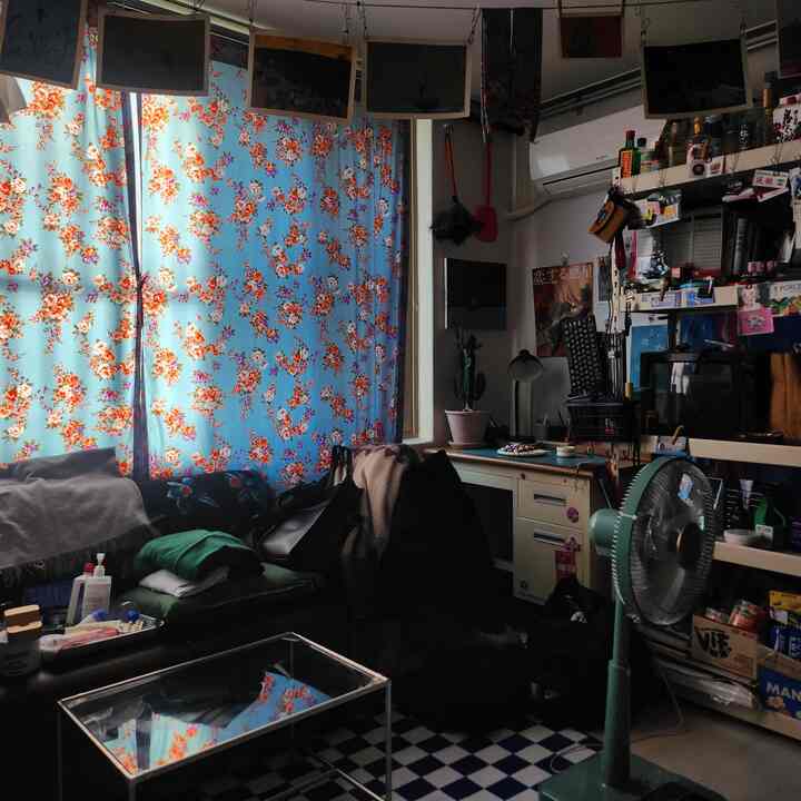 青い花柄のカーテンが特徴の狭いヴィンテージ風リビング、茶色のレザーソファとデスク、パントリー棚が配置された印象的な空間