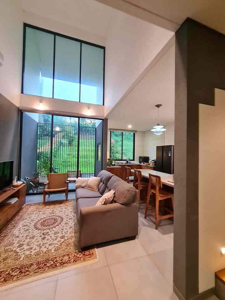 A modern natural living room featuring a gray sofa and wooden dining chairs, with large windows and high ceilings enhancing openness