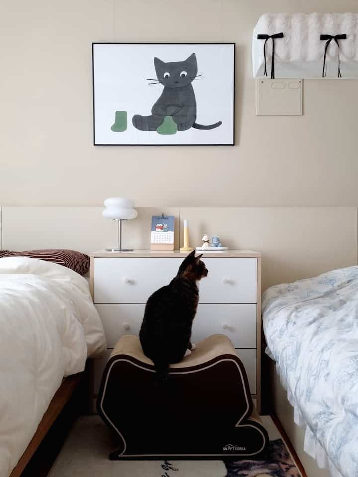 ベージュとホワイトの色調の寝室、中央に配置されたチェストと爪とぎの上に座る猫がいる雰囲気のあるインテリア