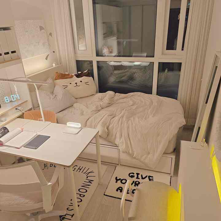 ホワイトとベージュのトーンの小さな寝室兼ホームオフィス、窓辺にベッドとデスクが配置された雰囲気のある空間