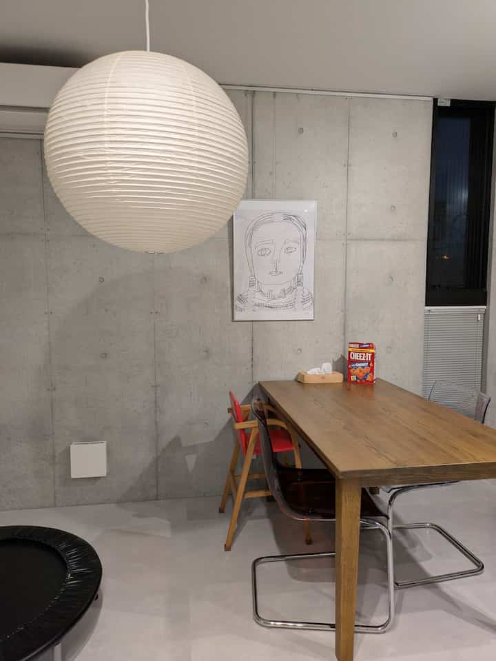 グレーと白を基調としたダイニングスペース、木製テーブルと透明な椅子、和風ペンダントライト、子供用ハイチェアとトランポリンが配置されたモダンなインテリア