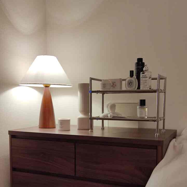 Brown and white toned 1K bedroom featuring a wooden table lamp and a stainless steel modular shelf with perfumes and candle, creating a cozy atmosphere