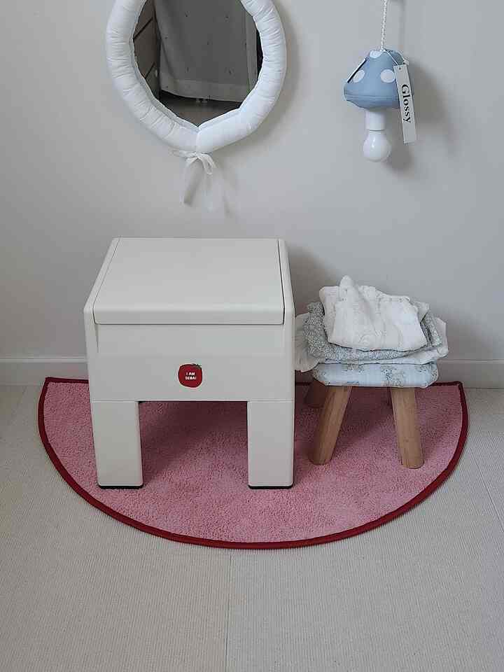 白とピンクの色調の子供部屋、白い収納スツールと小さな木製スツールが配置され、赤ちゃん用品が揃ったミニマルでナチュラルな空間