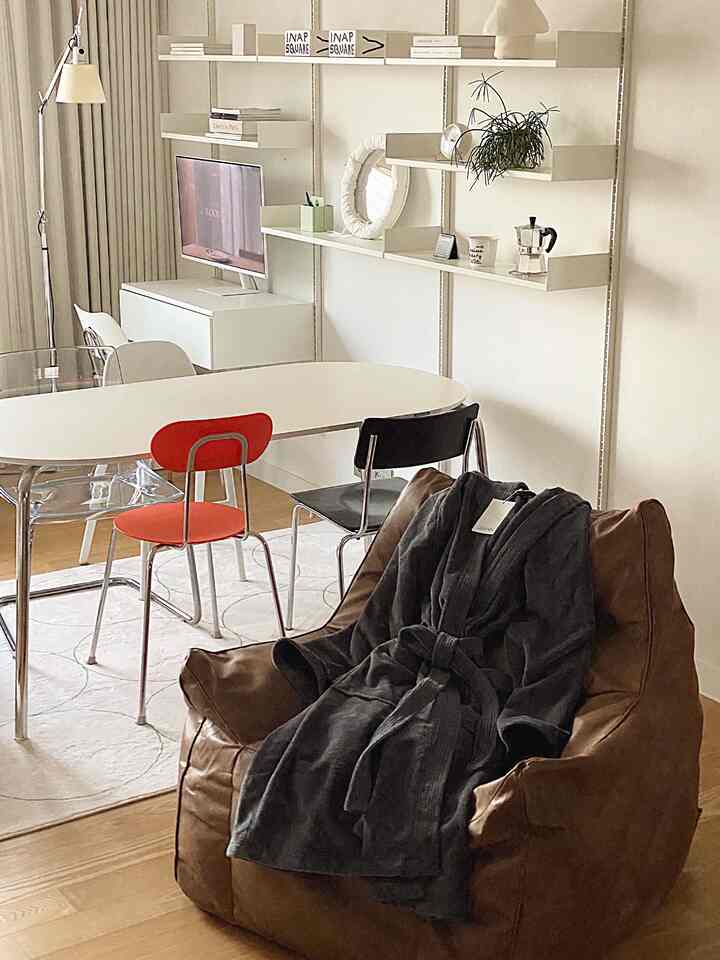 白と茶色のトーンのモダンなリビング兼ホームオフィス、壁掛け棚とビーンバッグソファ、楕円形のダイニングテーブルと様々な椅子が配置された空間