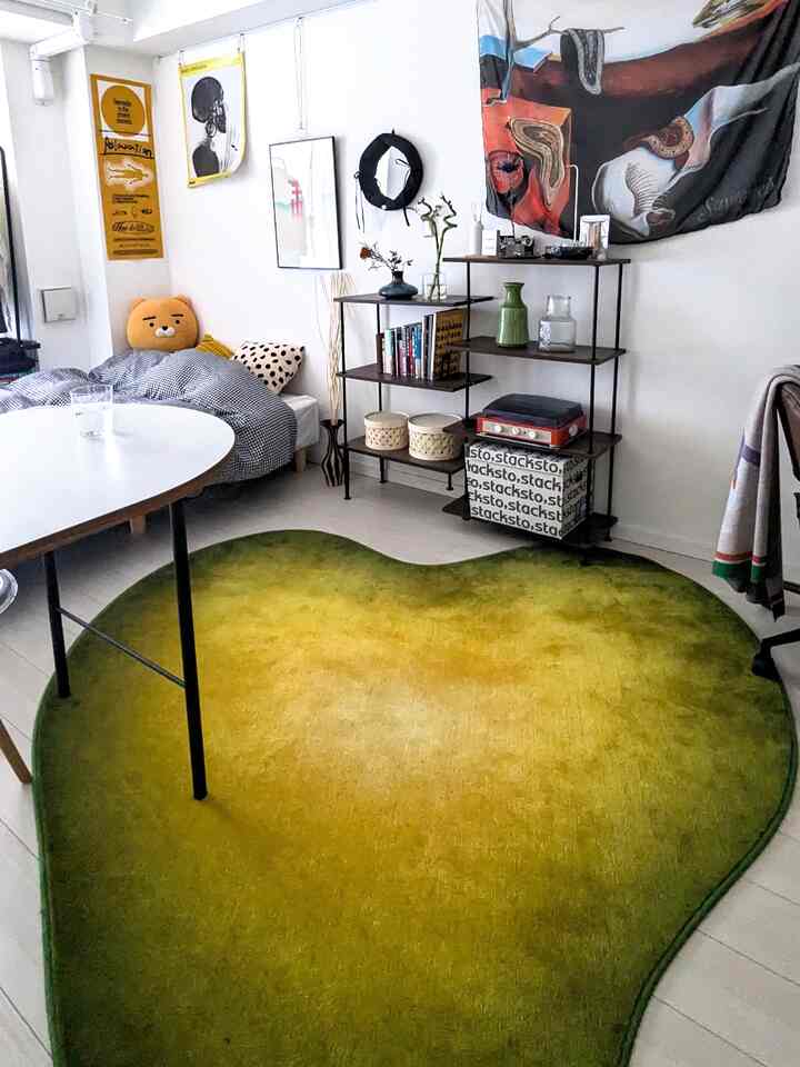 緑色調のワンルームリビング、チェック柄のベッドソファと個性的なラグ、花瓶や壁のポスターが配置された雰囲気ある空間