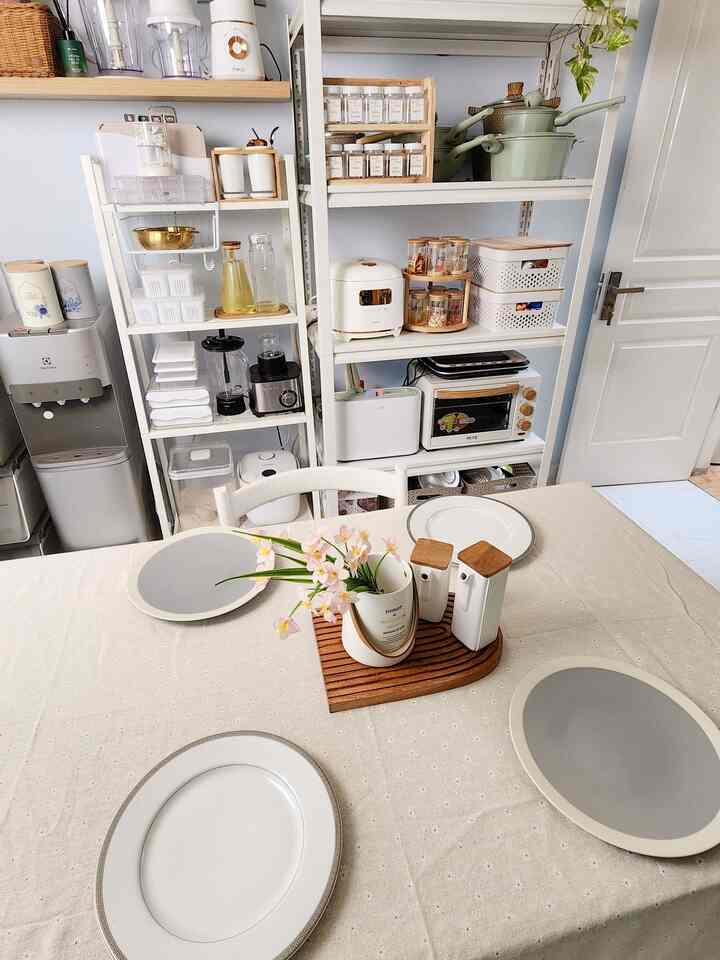 Bright white and beige toned dining room kitchen featuring a linen-covered table set with plates and organized storage shelves