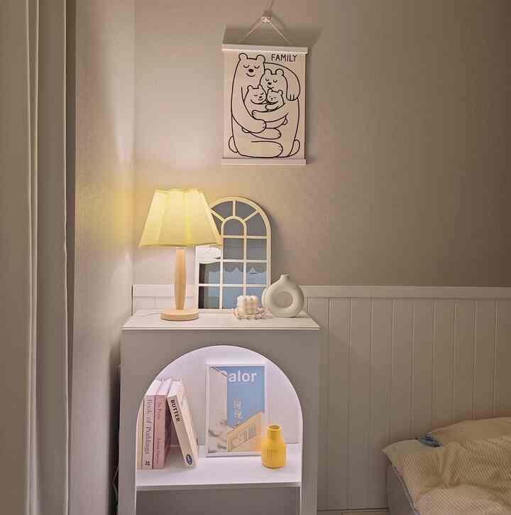温かみのあるベージュとホワイトの寝室、窓型の鏡とテーブルランプが置かれた収納台、家族の絵が配置された雰囲気のあるインテリア