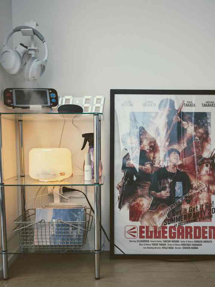 Bright white-walled studio space with transparent shelves holding white aroma diffuser and headphones, featuring a rock band poster on right, simple decor