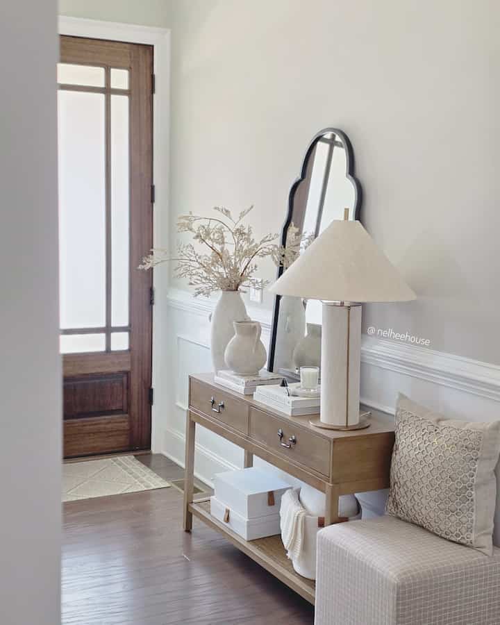 Ivory-toned entrance with wooden console table, large vase, table lamp, and cozy ottoman creating a natural and inviting atmosphere