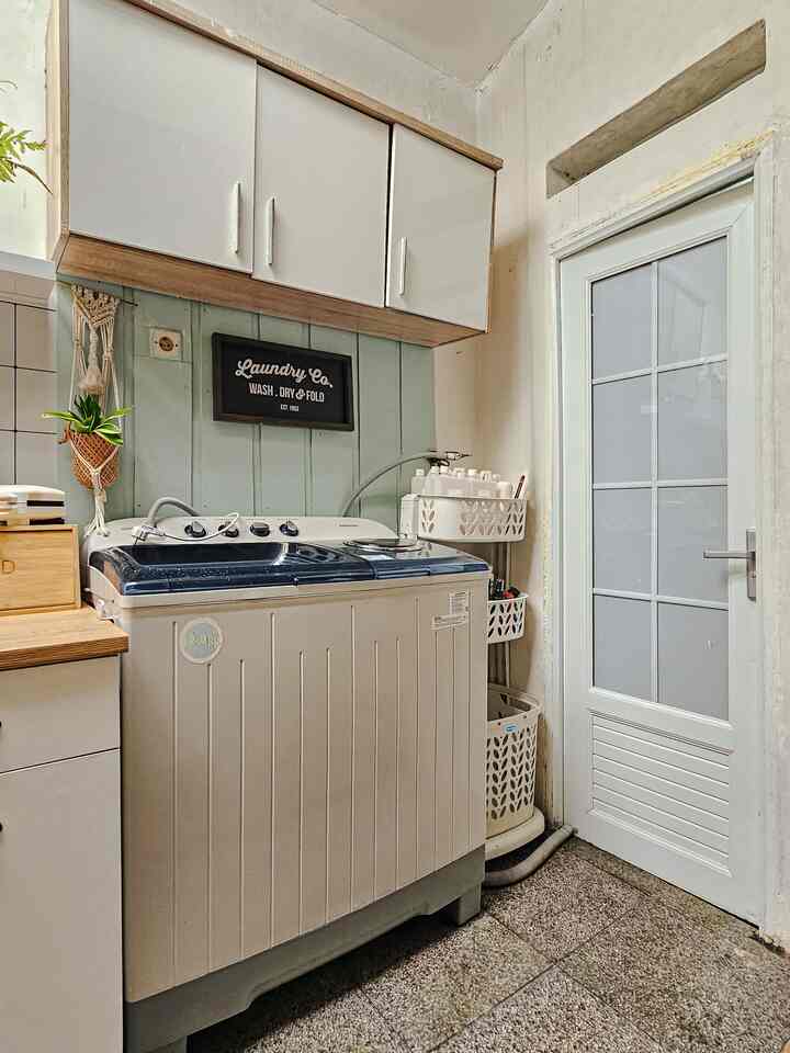 A narrow, vertical laundry space in white and mint tones featuring white cabinets, a washing machine, DIY wall battens, and a decorative plant for a cozy atmosphere