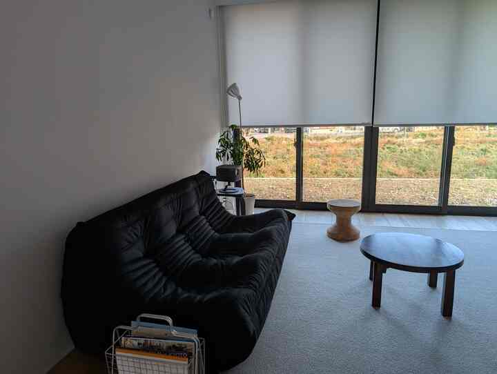 白い壁と大きな窓のあるリビング、黒いソファと丸いローテーブル、コルク製スツールが配置されたシンプルでナチュラルな空間