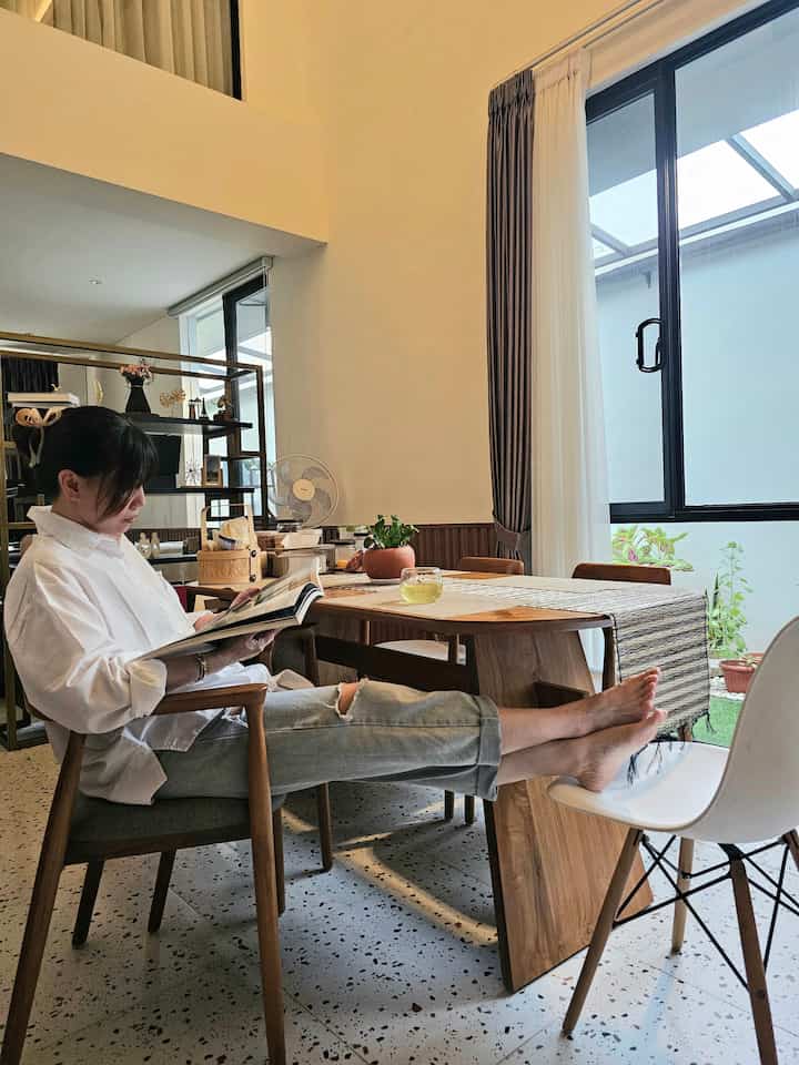 Natural wood-tone living dining room featuring a dining table beside a large window, with a person reading comfortably
